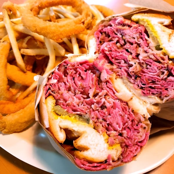 A large pastrami sandwich, cut in half, with a side of golden french fries and onion rings.